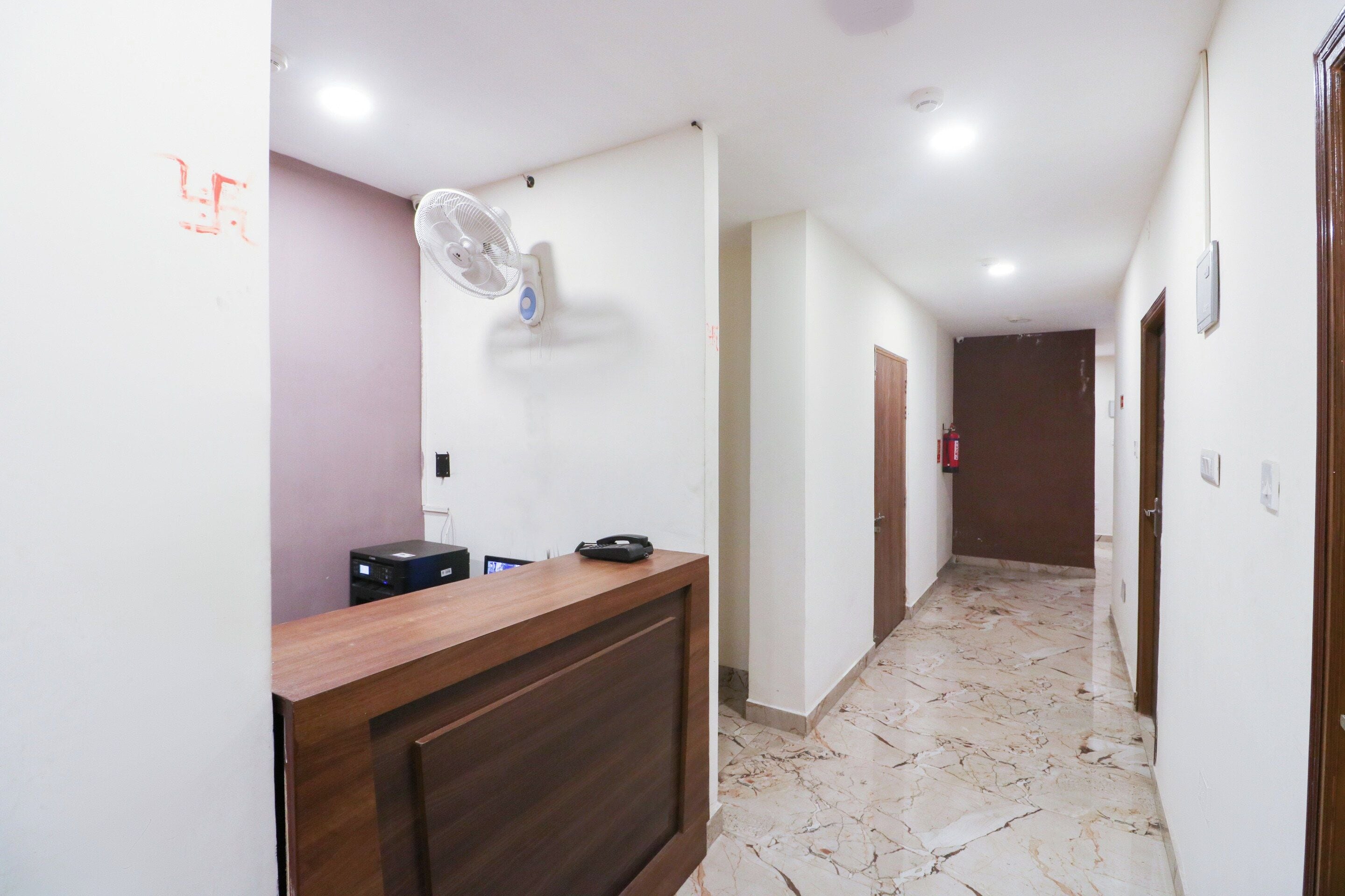 A hotel reception area with a wooden reception desk, a standing fan, a fire extinguisher, and a marble floor.