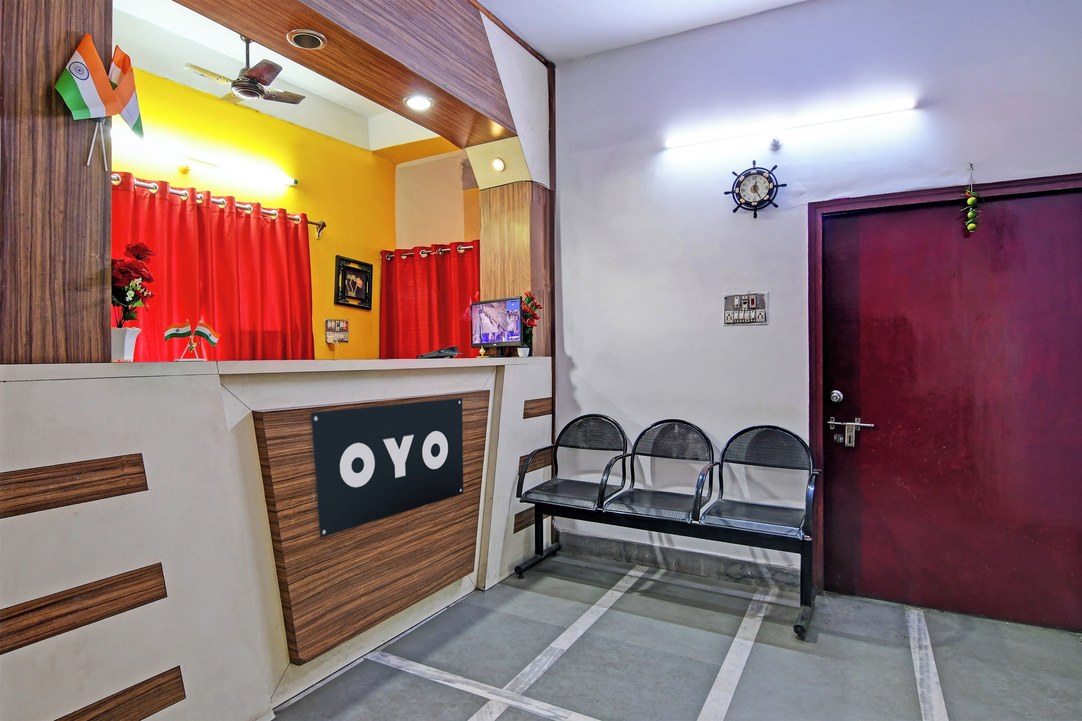 Un área de recepción con un mostrador de madera con la etiqueta 'OYO', una cortina roja, un ventilador de techo, un reloj en la pared y una puerta con manija.