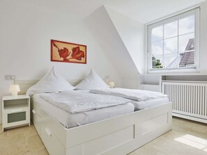 A bedroom with a bed, a nightstand, a lamp, a radiator, and a window with a view of buildings. - New Love - Suderstr. 18c (Sylt)
