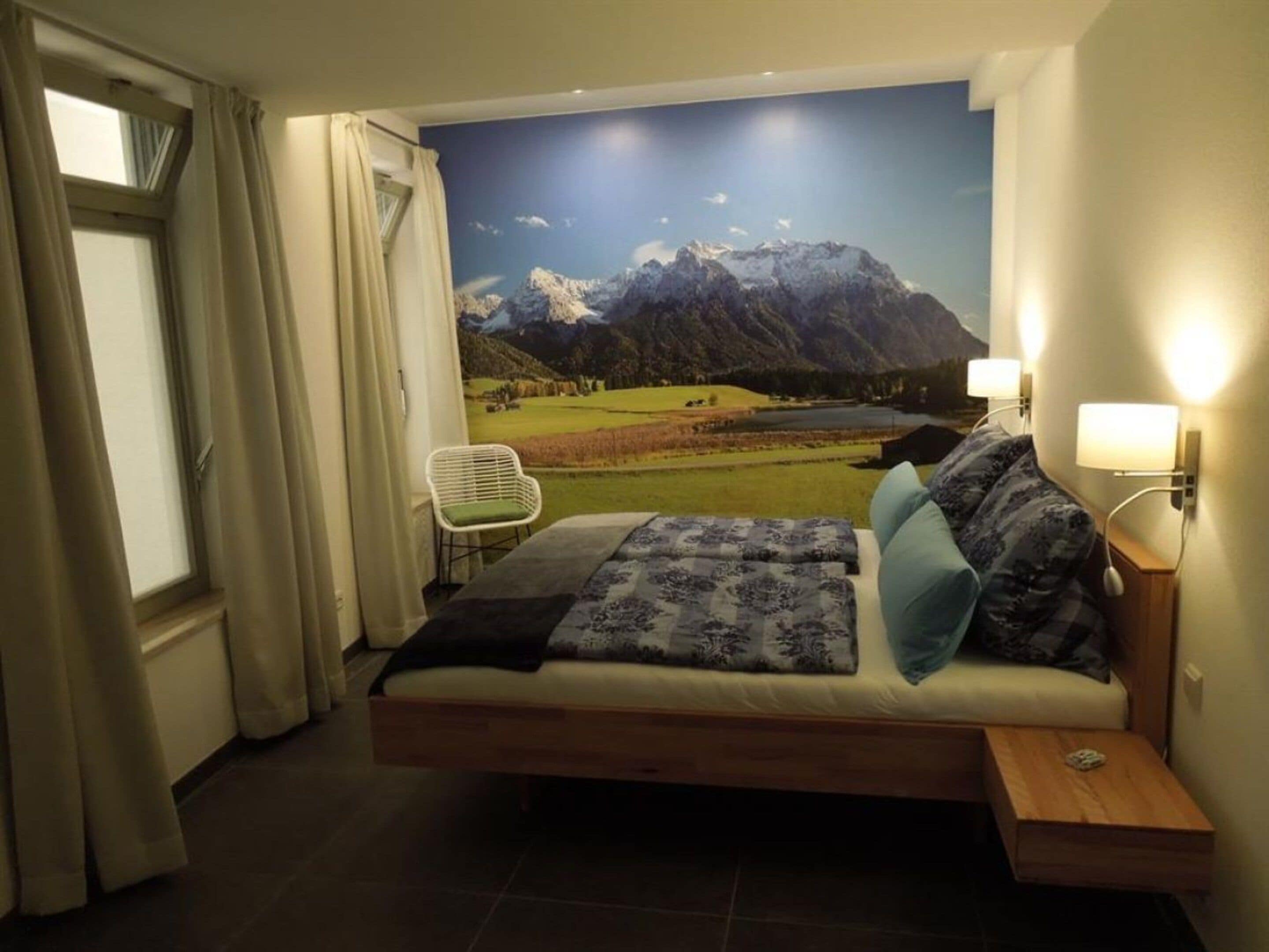 Un dormitorio con cama, una silla, una mesita de noche y un mural en la pared con un paisaje de montaña.