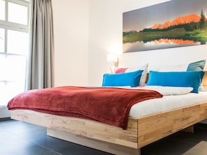 A bed with a red blanket, blue pillows, and a wooden headboard in a room with a large window and a painting of a landscape. - Holiday Apartment With 2 Bedrooms (Mittenwald)