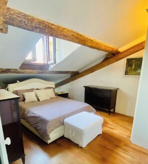 Ein Schlafzimmer mit schrägem Dach, einem Bett mit Kopfteil, einem Nachttisch, einer Kommode und einem Fenster.