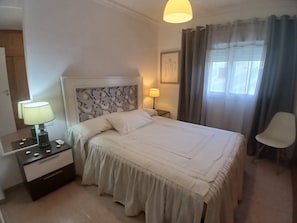 Une chambre d’hôtel bien rangée, avec un lit, des tables de chevet, une lampe, une chaise et une fenêtre avec des rideaux.