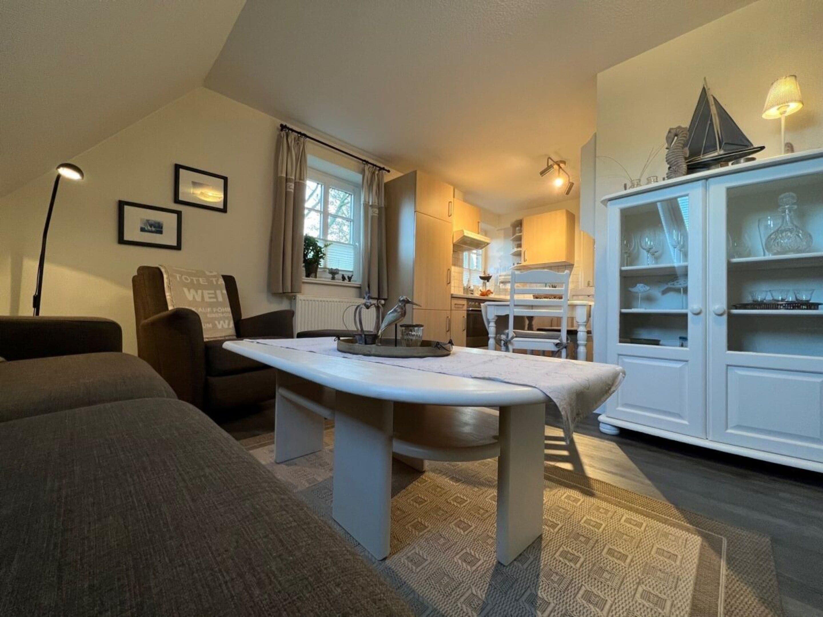 A cozy living room with a dining area, a kitchen in the background, and a white cabinet with glassware.