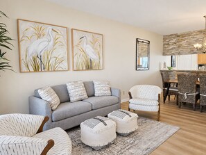 A living room with a grey sofa, patterned cushions, two white poufs, a patterned rug, and a dining area with wicker chairs and a wooden table. - Las Flores #B Spacious 2/2 W/Easy Beach Access (South Padre Island)