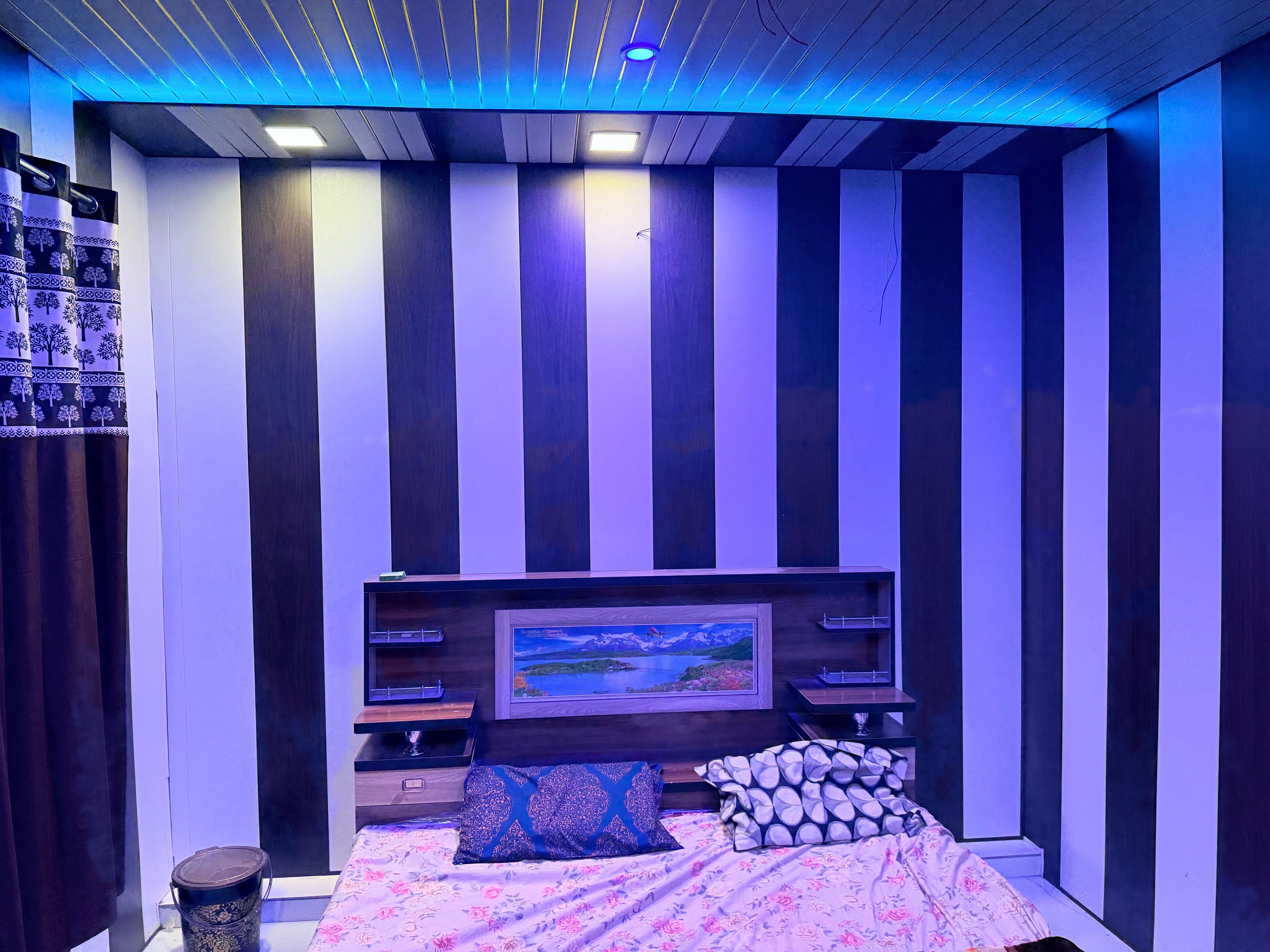 Un dormitorio con cama, mesita de noche, televisor montado en la pared y repisas con objetos decorativos.