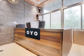 Un mostrador de recepción con un cartel que dice 'OYO', una computadora portátil, un ventilador y una ventana con vista a árboles.