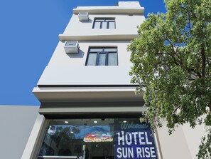 Ein Hotel Eingang mit blauem Himmel, einem Schild mit der Aufschrift 'Welcome to Hotel Sun Rise' und einer Klimaanlage an der Fassade.