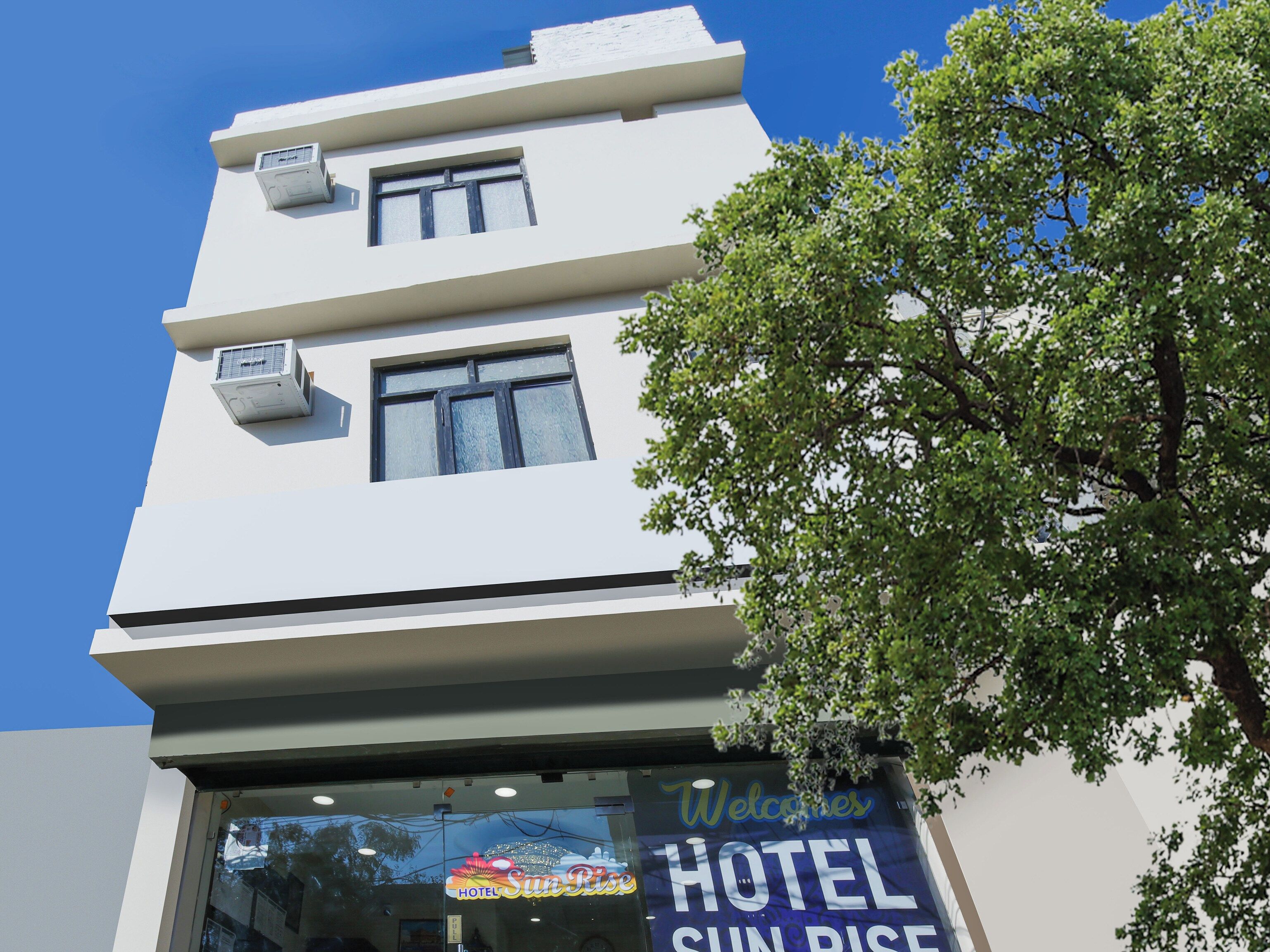 Un hotel con cielo despejado, un cartel que dice 'Welcome to HOTEL Sun Rise' y un árbol en primer plano.