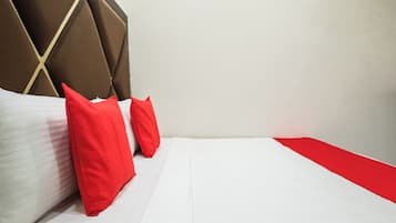Un lit bien fait, avec du linge de lit blanc et deux oreillers rouges, une tête de lit recouverte de tissu marron et un élément décoratif rouge sur le côté du lit.
