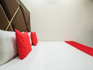 Ein ordentlich bezogenes Bett mit weißen Leinentüchern und zwei roten Kissen, ein braunes Polsterkopfteil und ein roter Akzent am Bettrand.