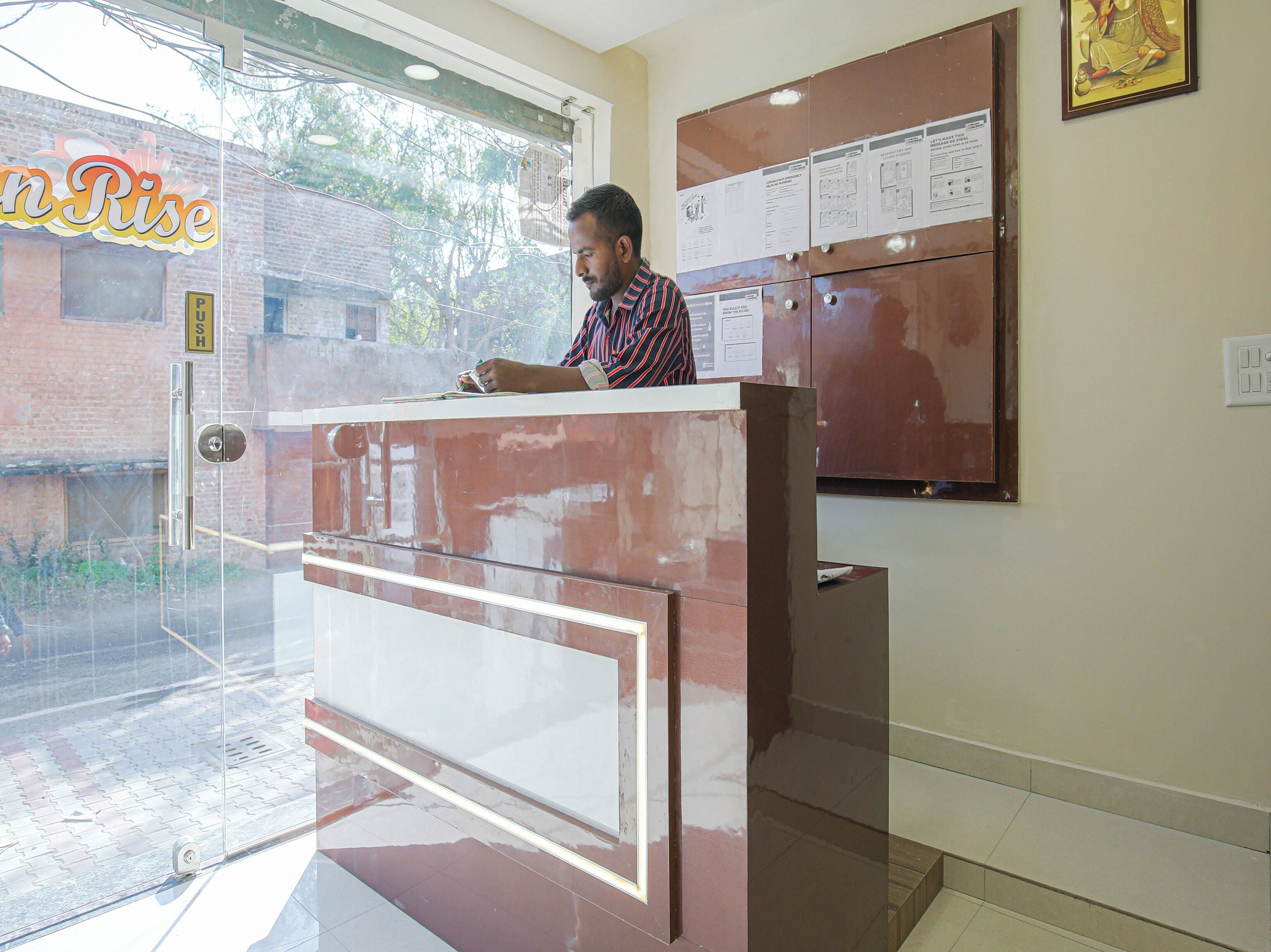 A reception desk with a person seated behind it, a bulletin board with notices, and a window with a sign reading 'SunRise'.