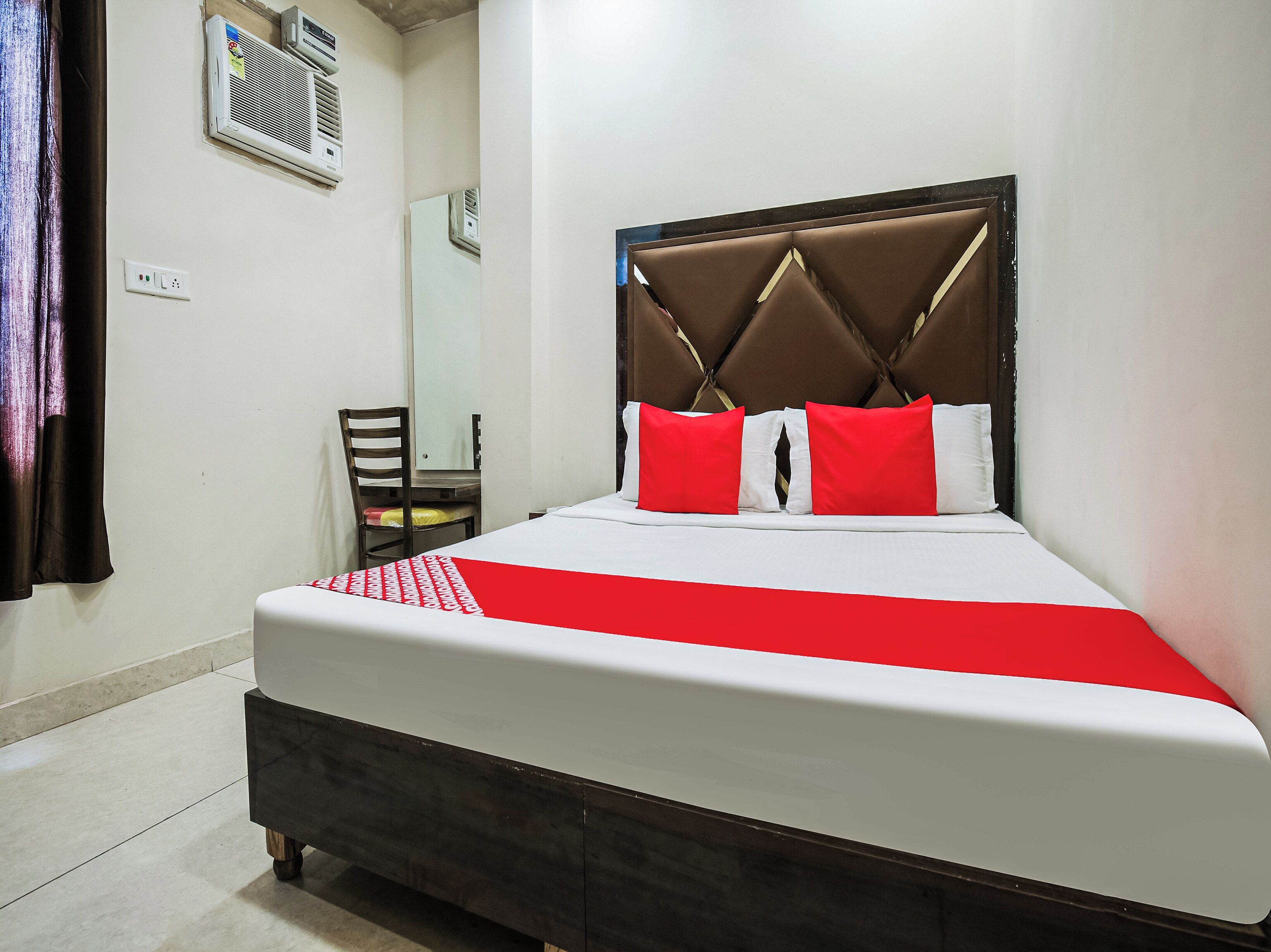 A hotel room with a bed, a chair, a mirror, and an air conditioning unit.