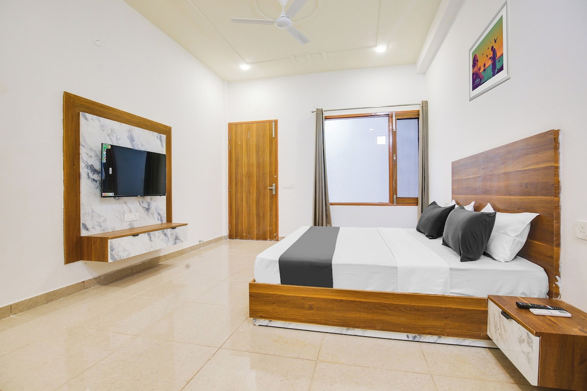 A bedroom with a bed, a TV mounted on a marble wall, a wooden headboard, a nightstand, and a window with curtains.