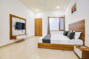 A bedroom with a bed, a TV mounted on a marble wall, a wooden headboard, a nightstand, and a window with curtains. - Hotel O by OYO GRAND STAY (Ludhiana)
