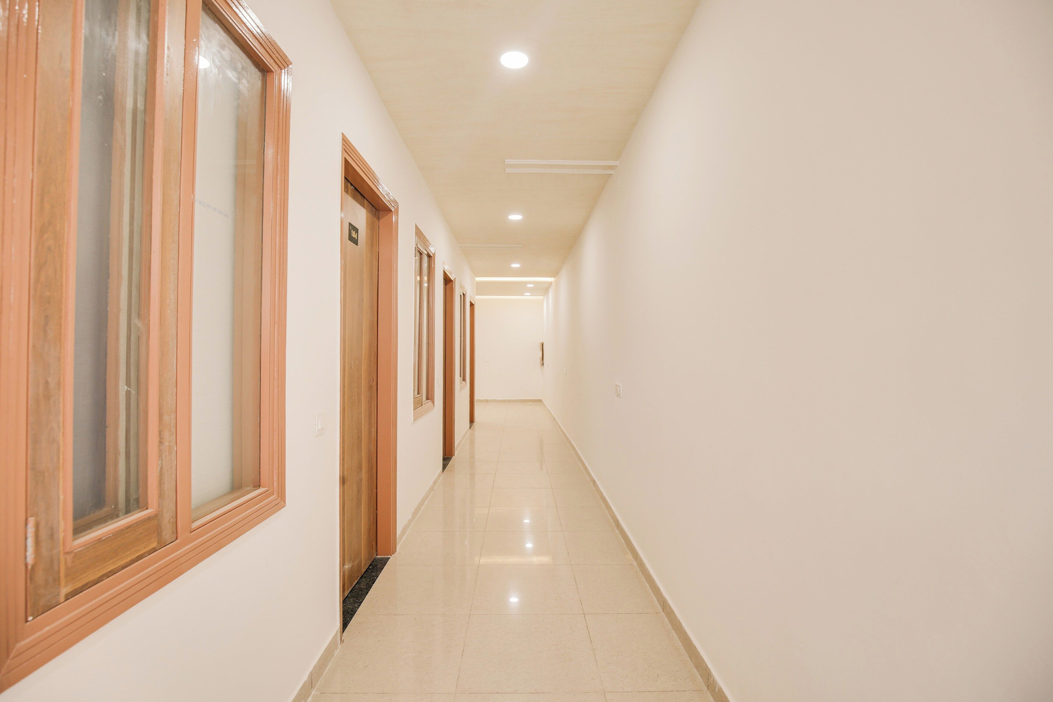 Un pasillo largo y estrecho con puertas y ventanas de madera en un lado, que conduce a una habitación luminosa con suelo de baldosas.