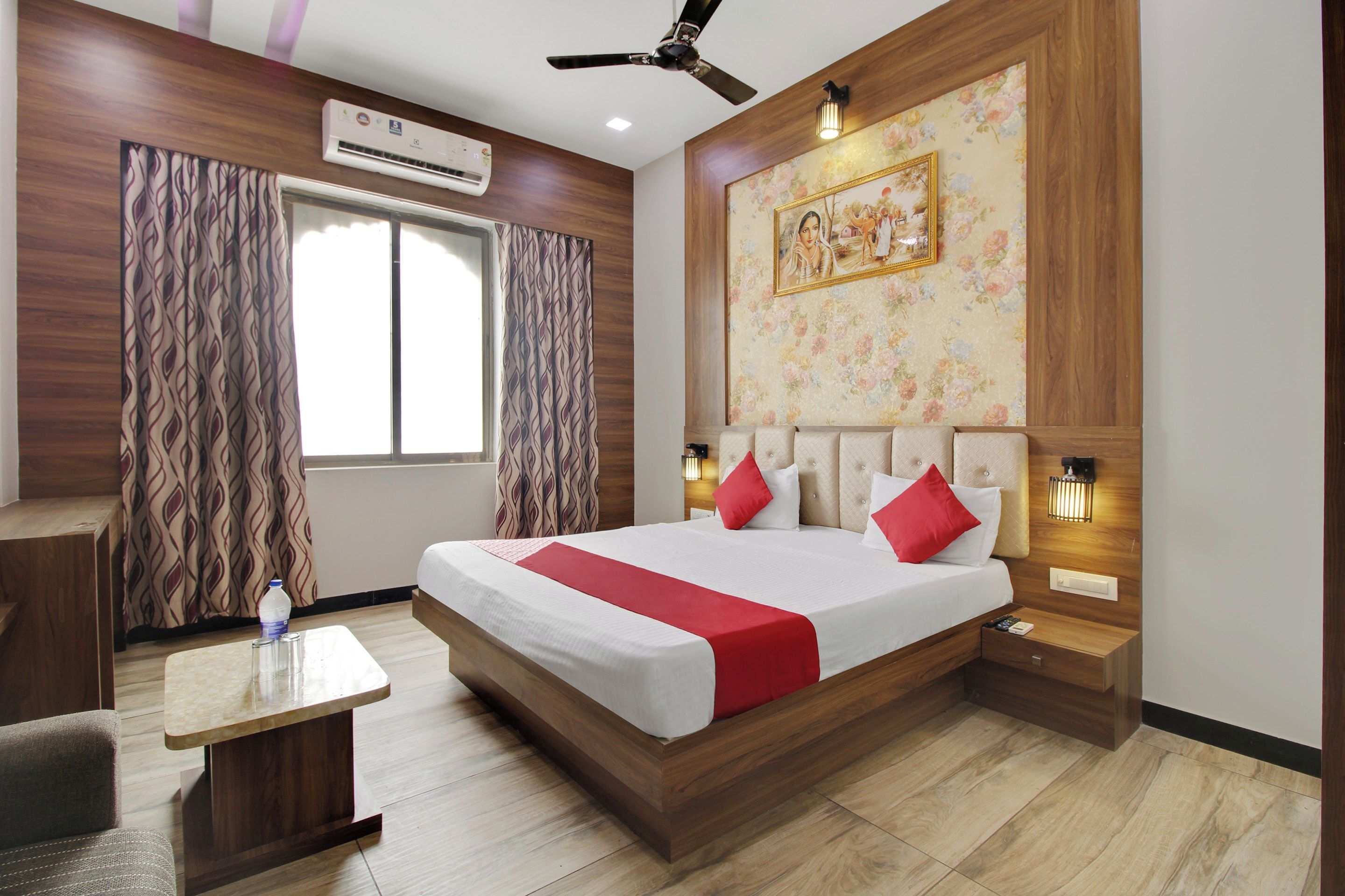A hotel room with a bed, a desk, a chair, a window with curtains, an air conditioning unit, a ceiling fan, a wall-mounted light, and a framed picture.