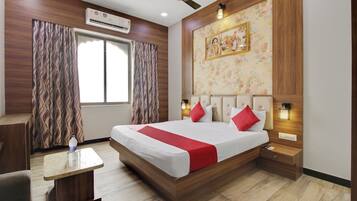 A hotel room with a bed, a desk, a chair, a window with curtains, an air conditioning unit, a ceiling fan, a wall-mounted light, and a framed picture.