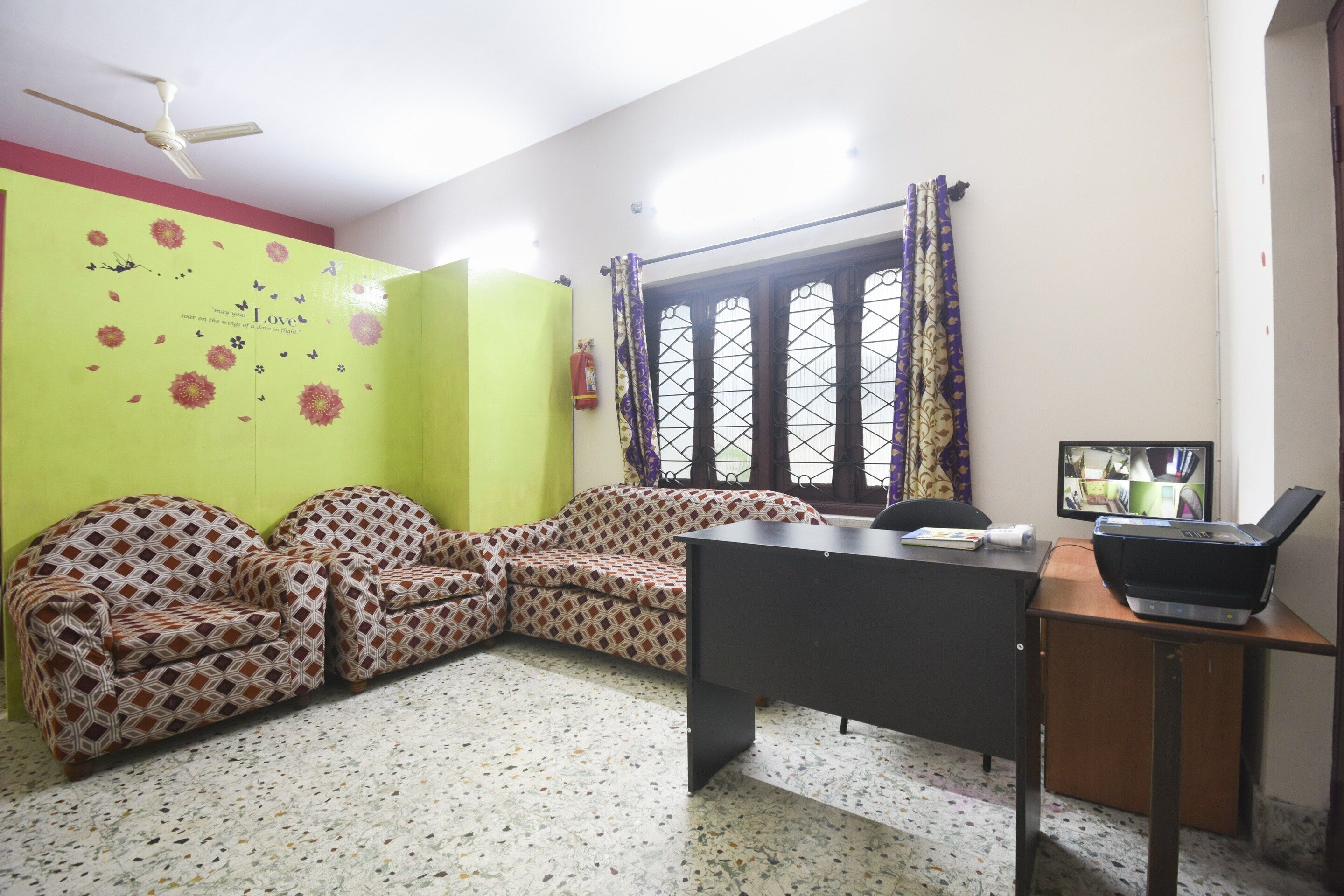 A room with a desk, a patterned sofa, a window with curtains, and a ceiling fan.