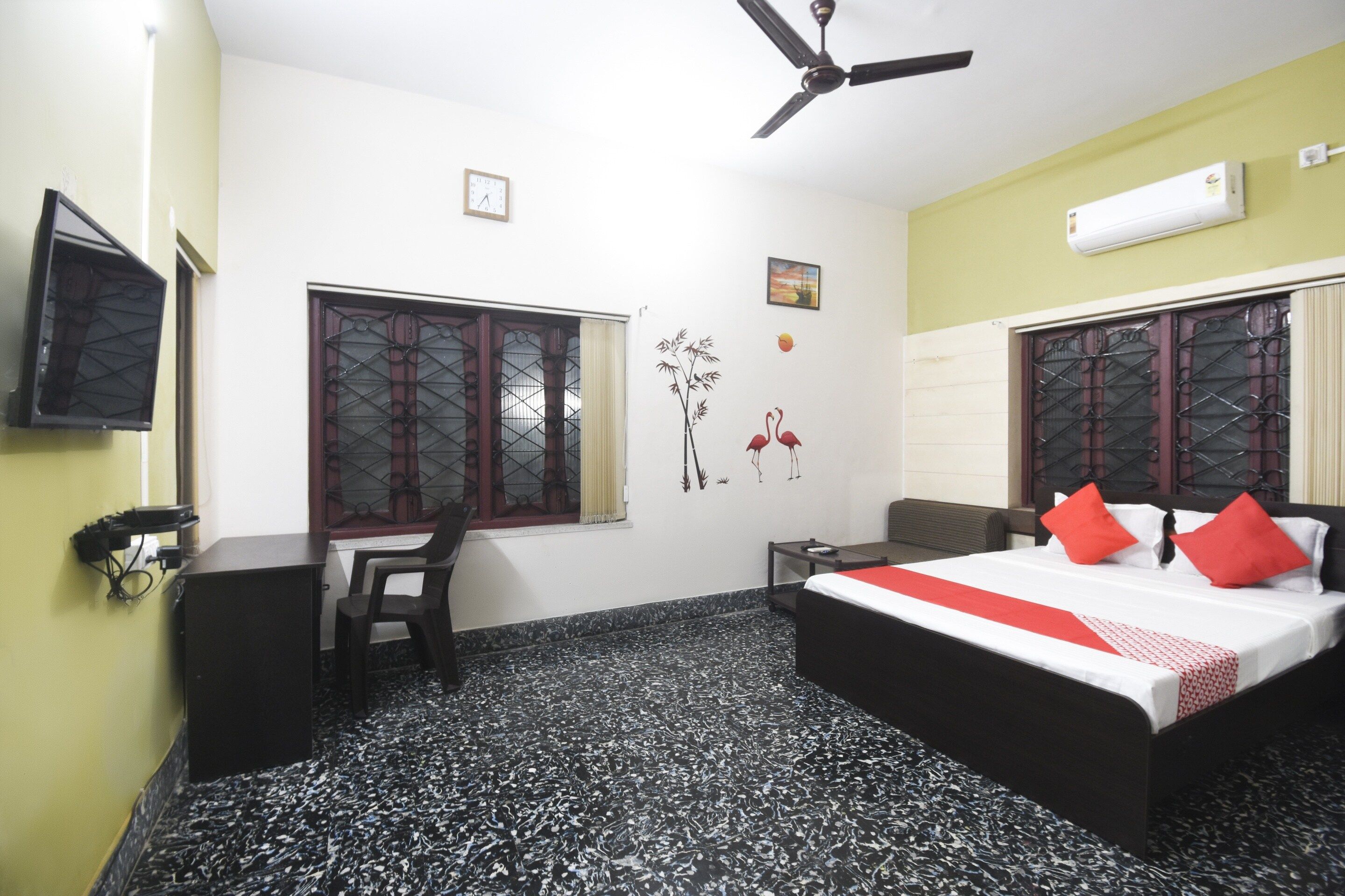 Habitación de hotel con cama, escritorio, silla, ventilador de techo y televisor montado en la pared.
