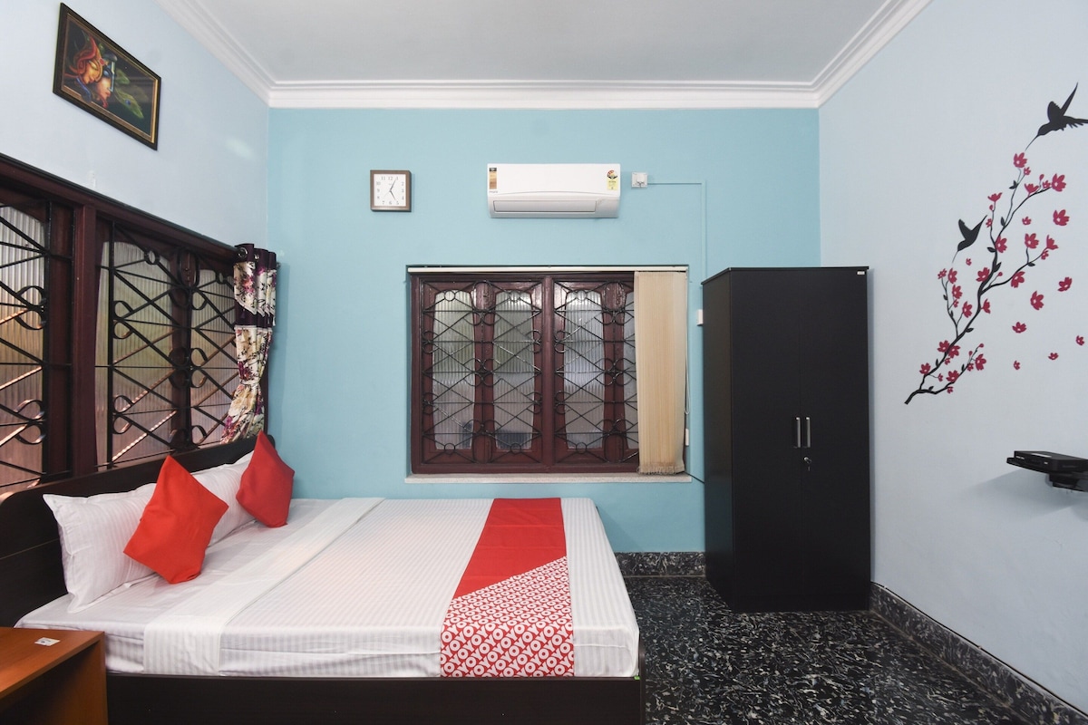 A hotel room with a bed, a wardrobe, a window with a metal grill, an air conditioning unit, and a wall decoration of a bird and flowers.