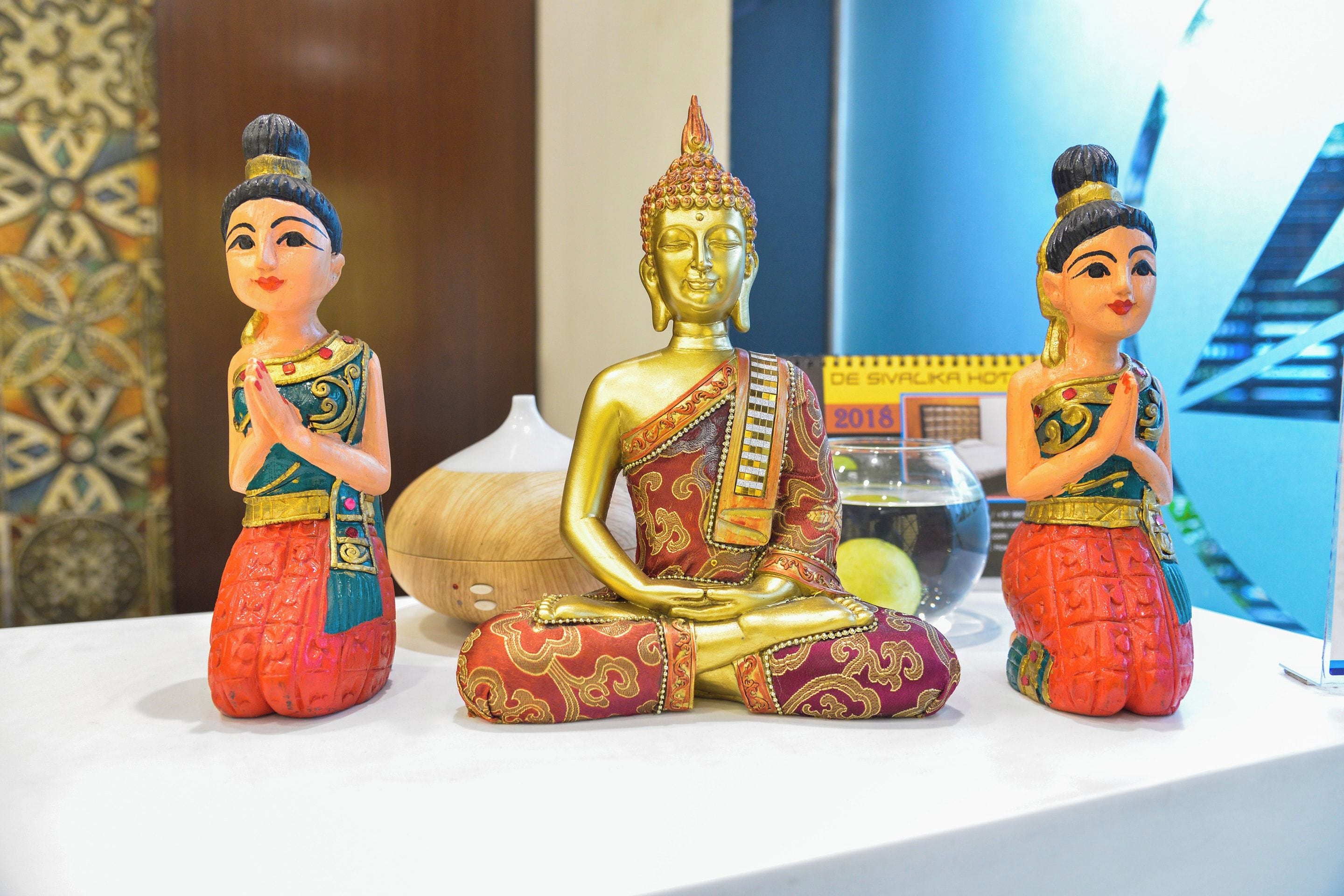 Three statues of a seated figure in traditional attire, a white vase, and a blue patterned wall.