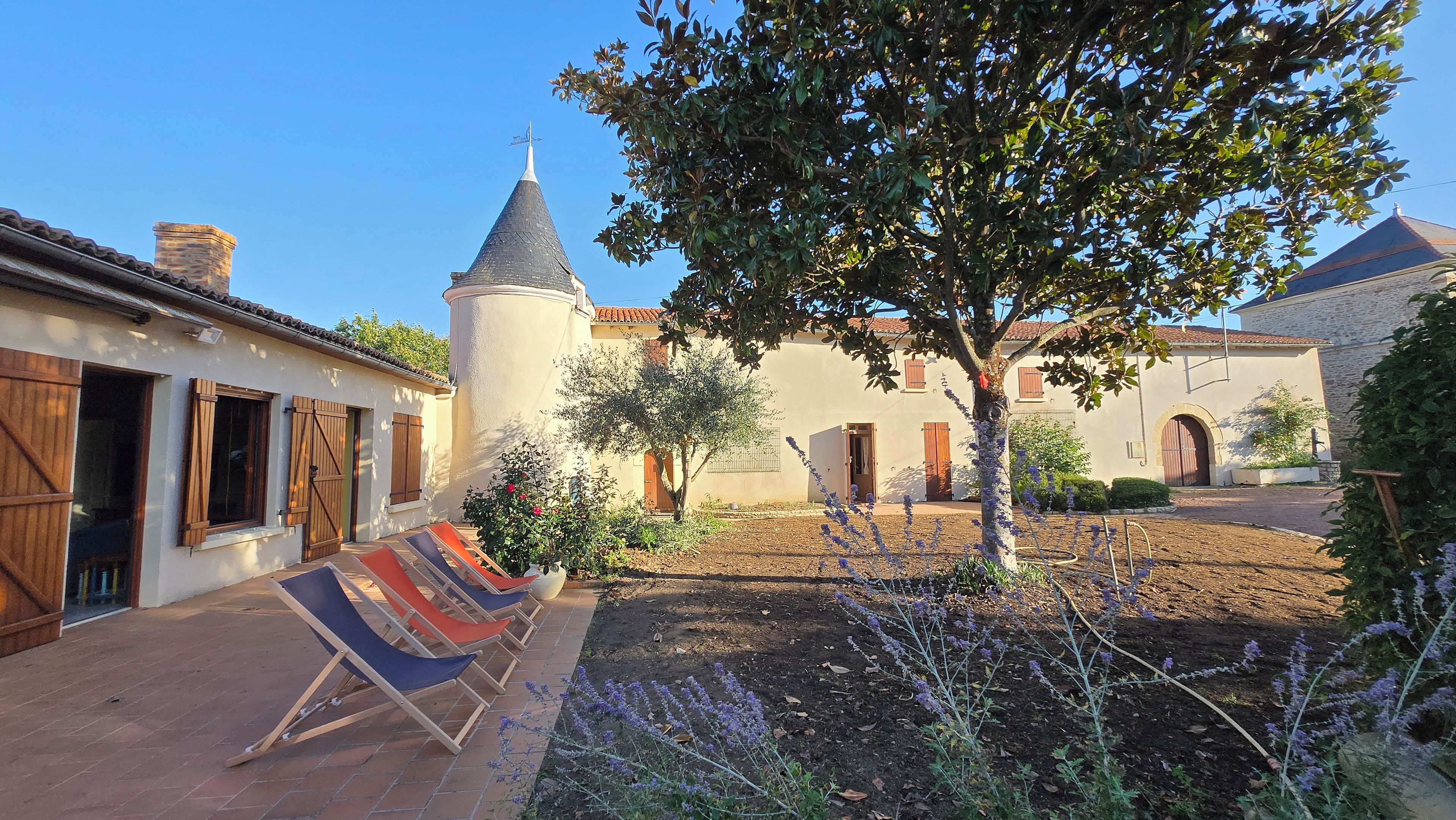 A courtyard with a building featuring a conical roof, a tree, and a row of colorful sun loungers.