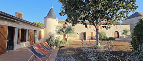 A courtyard with a building featuring a conical roof, a tree, and a row of colorful sun loungers.