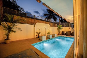 An outdoor swimming pool with a tiled edge, surrounded by potted plants and a fenced area with a view of palm trees. - Staymaster White Castle - 7BHK Villa (Goa)