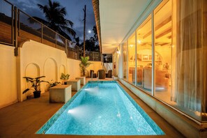 A swimming pool with a tiled edge, a covered seating area with chairs, and a view of the adjacent building. - Staymaster White Castle - 7BHK Villa (Goa)