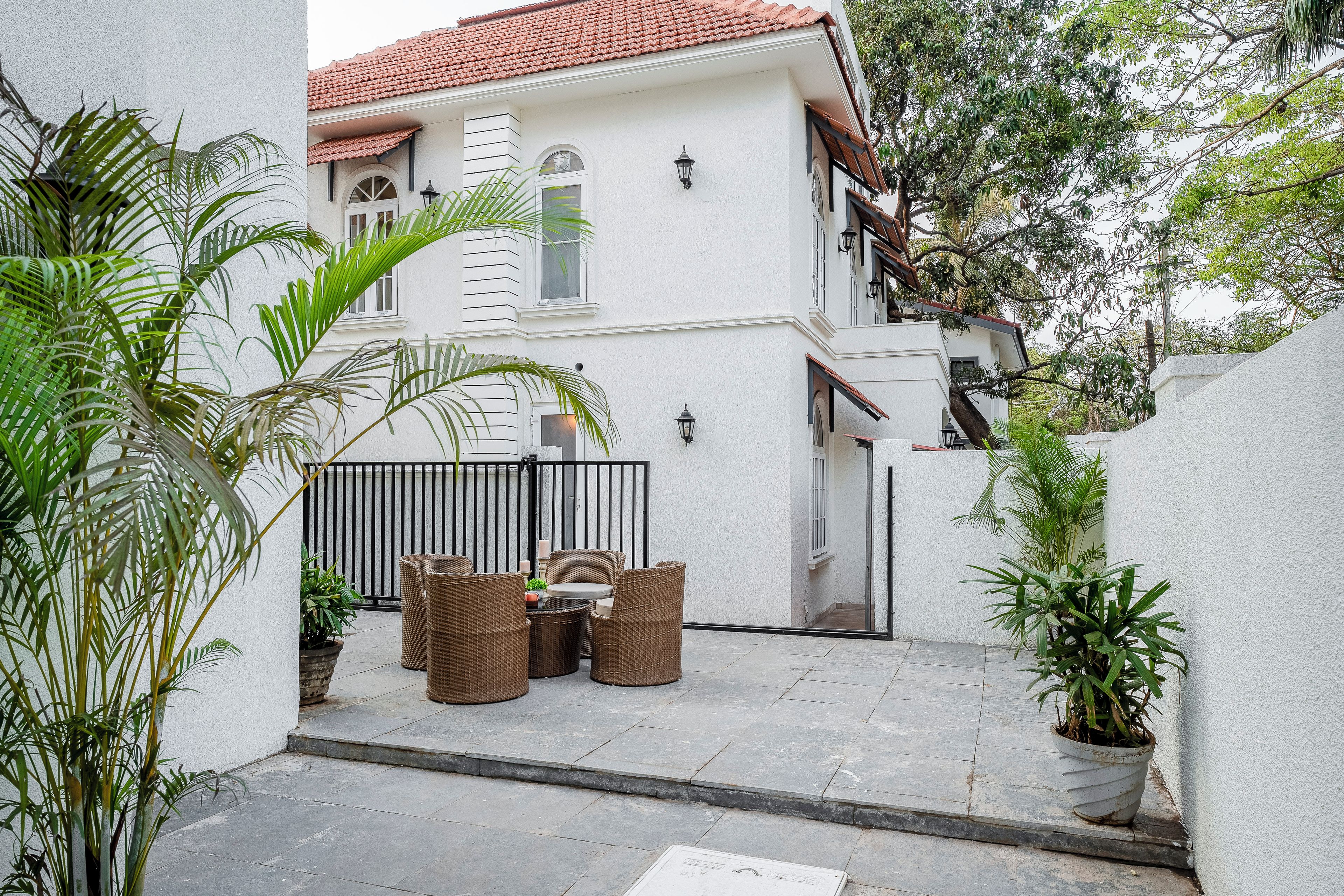 A white building with a tiled roof, a gated patio area with wicker furniture, and potted plants.