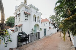 A white building with a red-tiled roof, a gated entrance, and a dirt path leading to it. - Staymaster White Castle - 7BHK Villa (Goa)