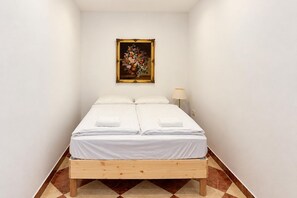 A single bed with white linens and a wooden frame, a nightstand with a lamp, and a floral painting on the wall. - A Little Vienna for Two near Westbahnhof (Vienna)