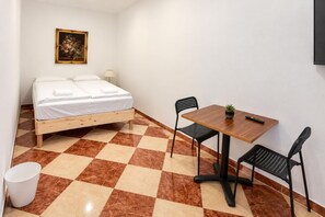 Ein Zimmer mit einem Bett, einem Tisch mit Stühlen, einer kleinen Topfpflanze und einem Bild an der Wand.