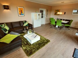 Una sala de estar con un sofá marrón, una mesa de centro blanca y un tapete verde. Hay un comedor con sillas verdes y una mesa blanca.