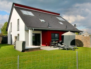 Una casa moderna de dos pisos con techo inclinado, un patio con mobiliario exterior y un muro de piedra.