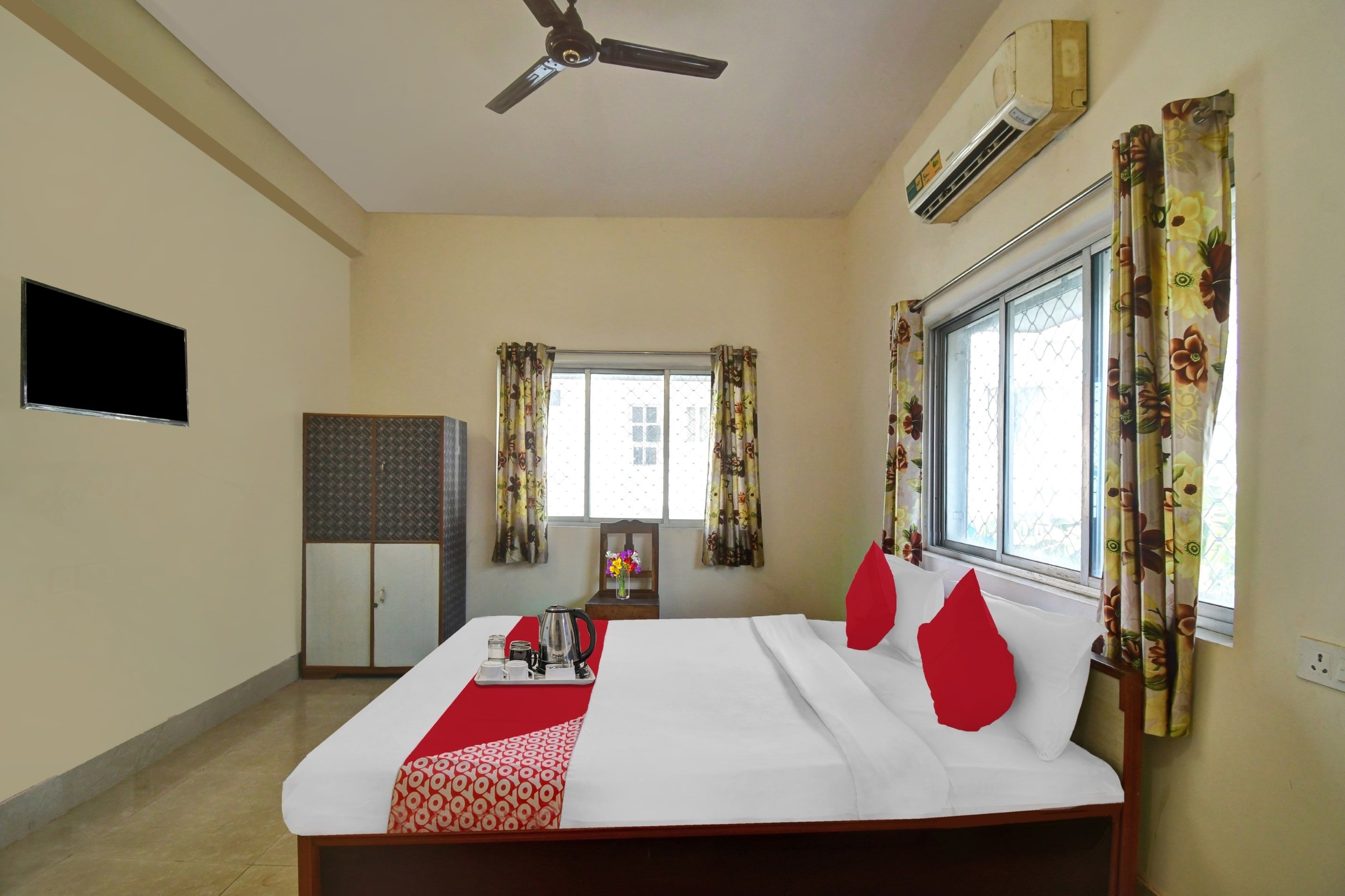 Habitación de hotel con cama, ventilador de techo, aire acondicionado, televisor, armario, ventana con cortinas y una mesa con mantel rojo.