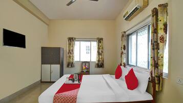 Habitación de hotel con cama, ventilador de techo, aire acondicionado, televisor, armario, ventana con cortinas y una mesa con mantel rojo.