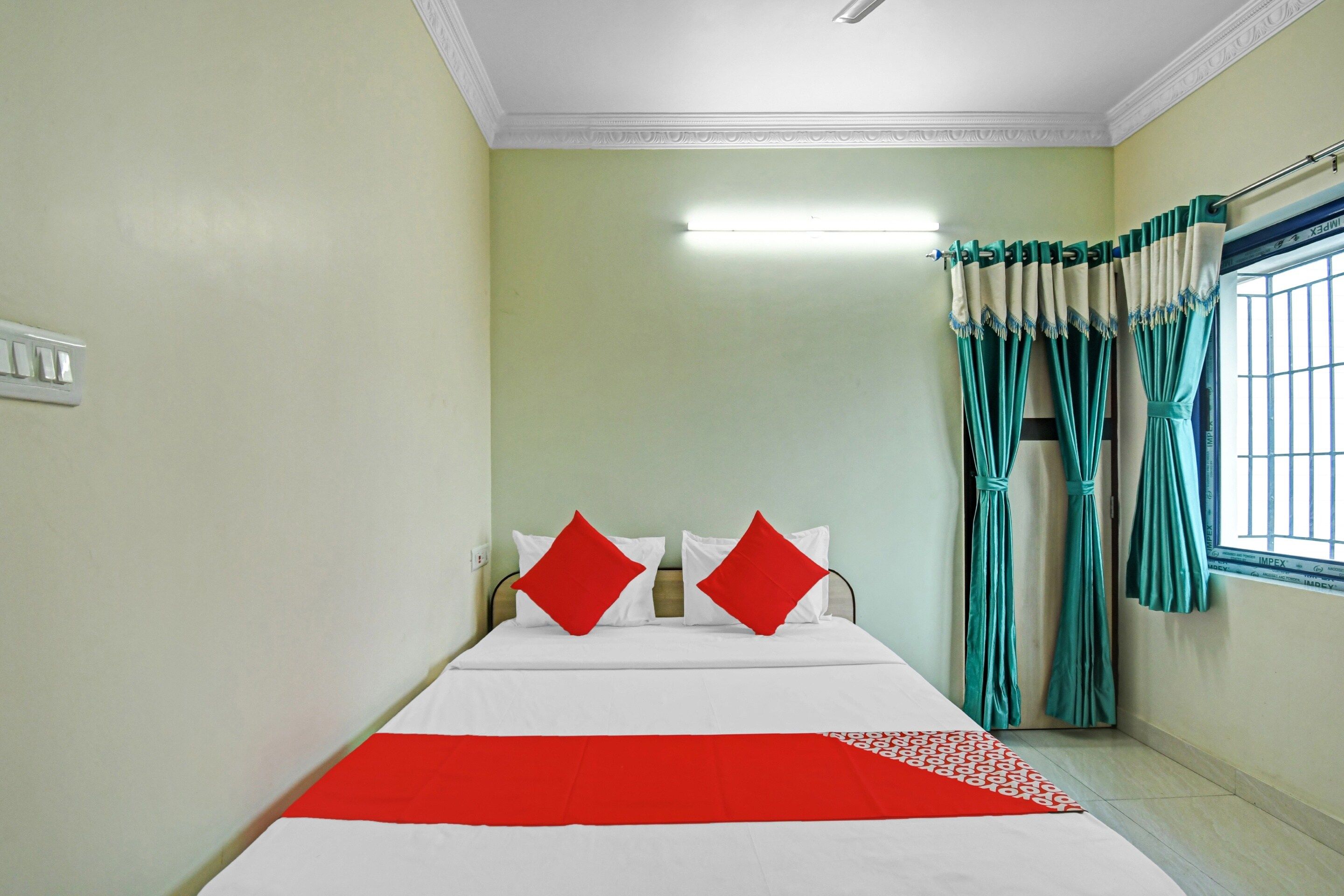 A hotel room with a bed, two red pillows, a white and red bedspread, a window with green curtains, and a light fixture on the ceiling.
