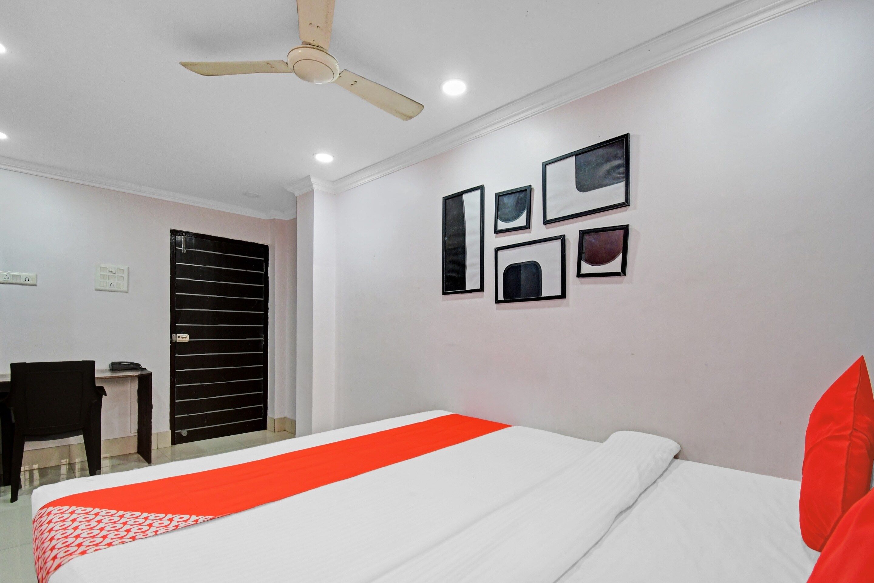 Habitación de hotel con cama, escritorio, silla, ventilador de techo y cuadros en la pared.