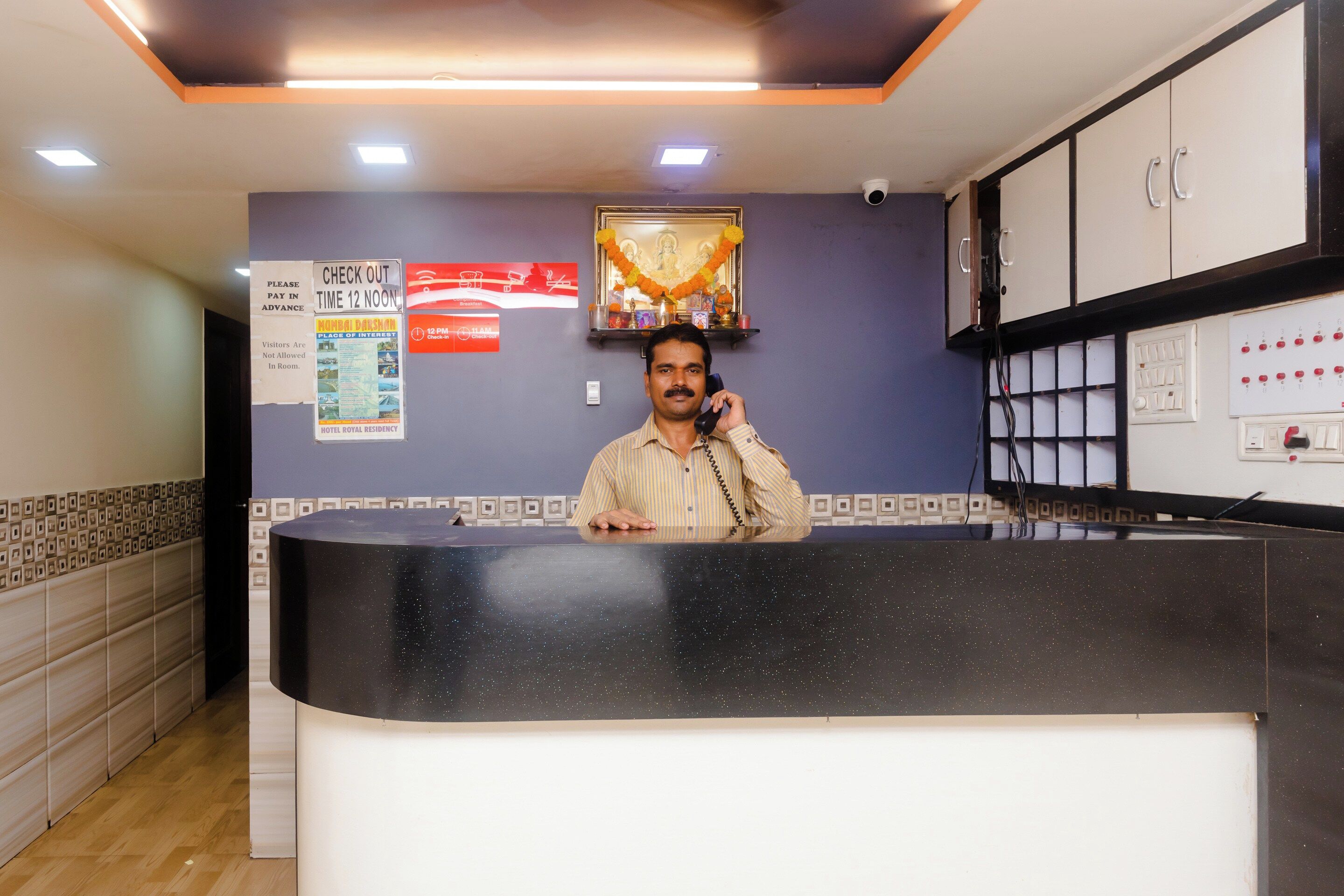 A hotel reception desk with a man standing behind it, a check-out sign, and a wall-mounted security camera.