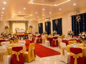 Un salón de banquetes con mesas redondas, sillas con cintas doradas y un escenario con decoración floral.