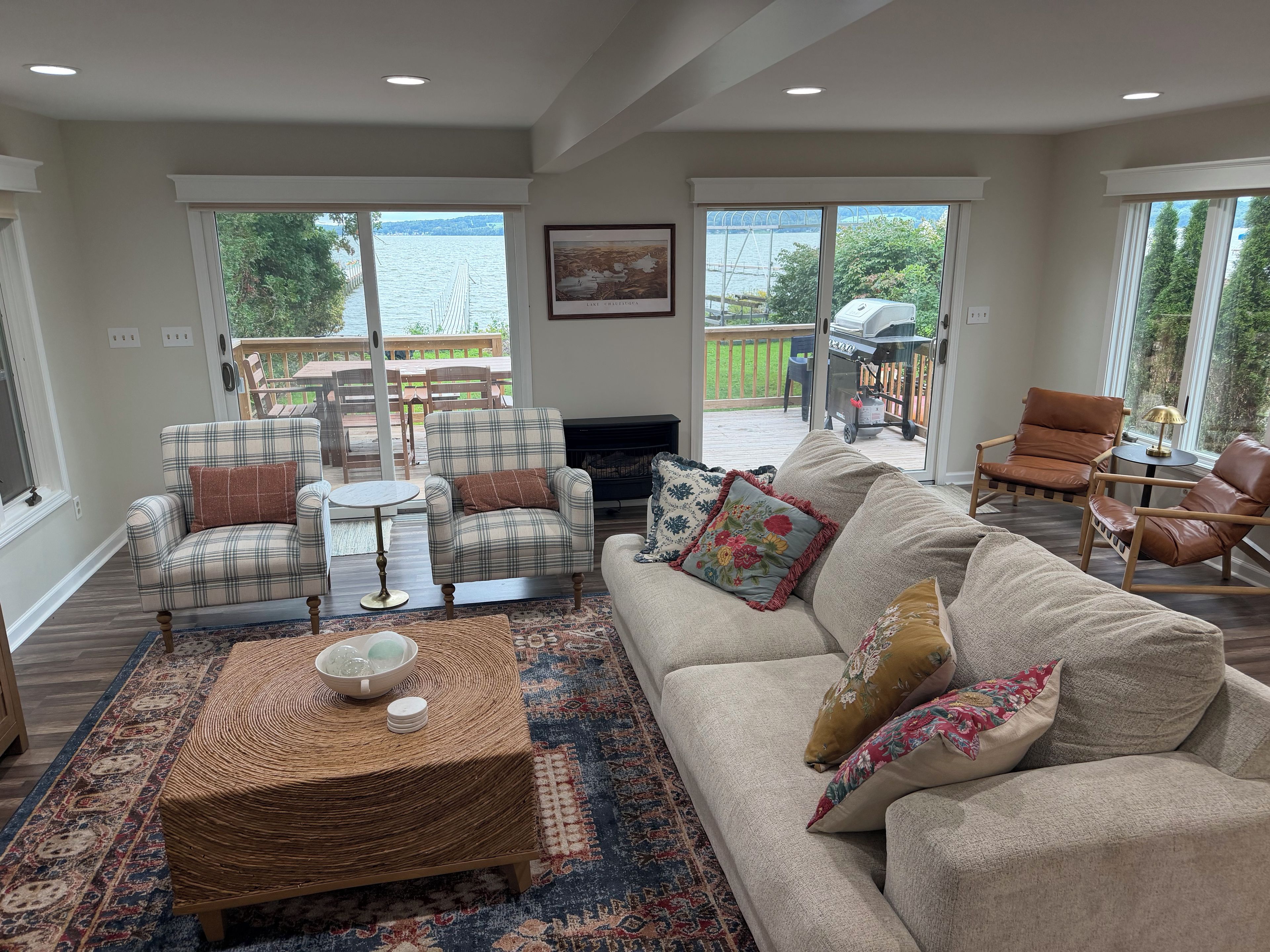 A living room with a sofa, armchairs, a coffee table, and a fireplace. There are large windows offering a view of a deck and outdoor area.