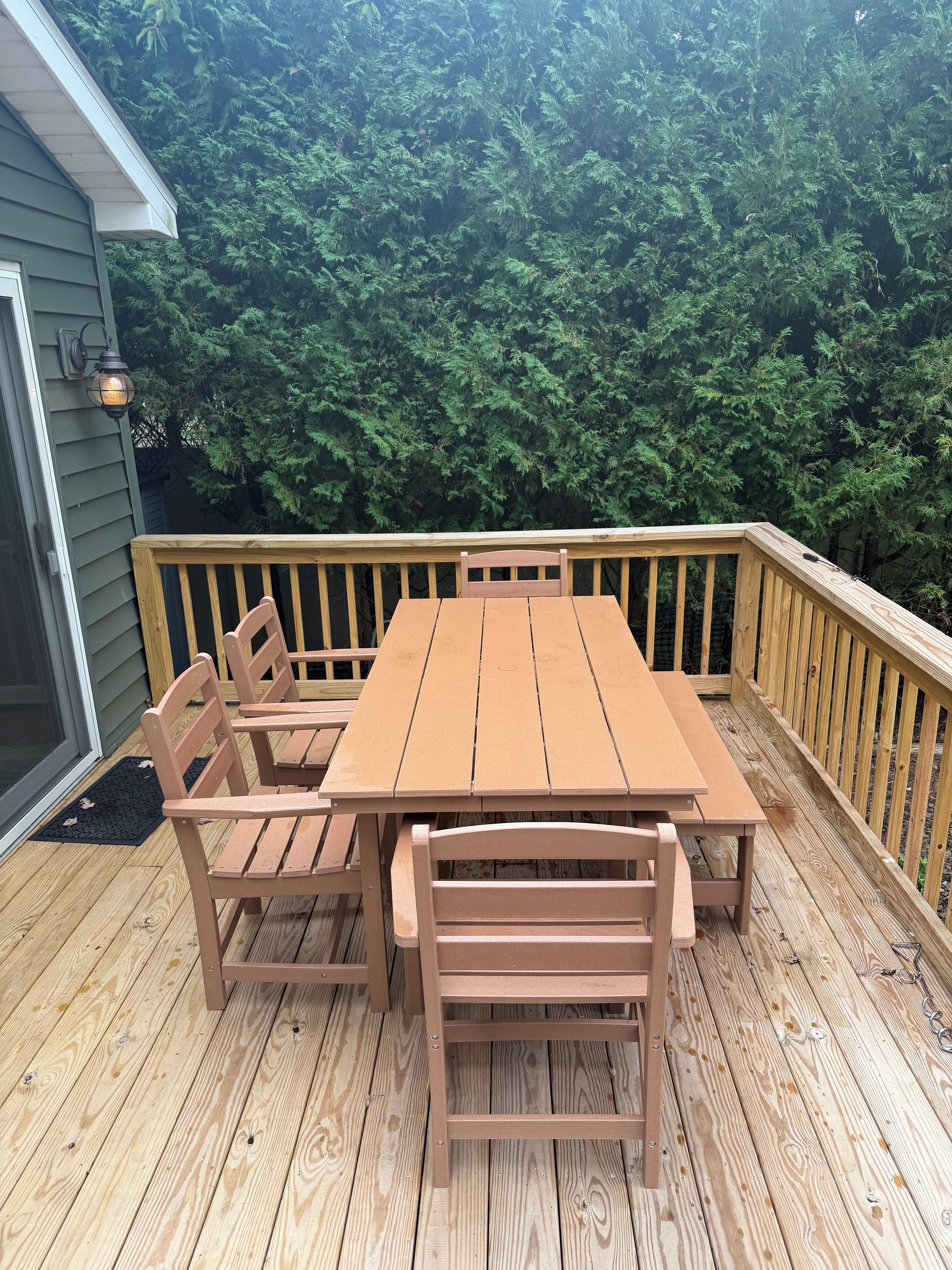 A wooden deck with a table and chairs, surrounded by a railing and dense foliage in the background.