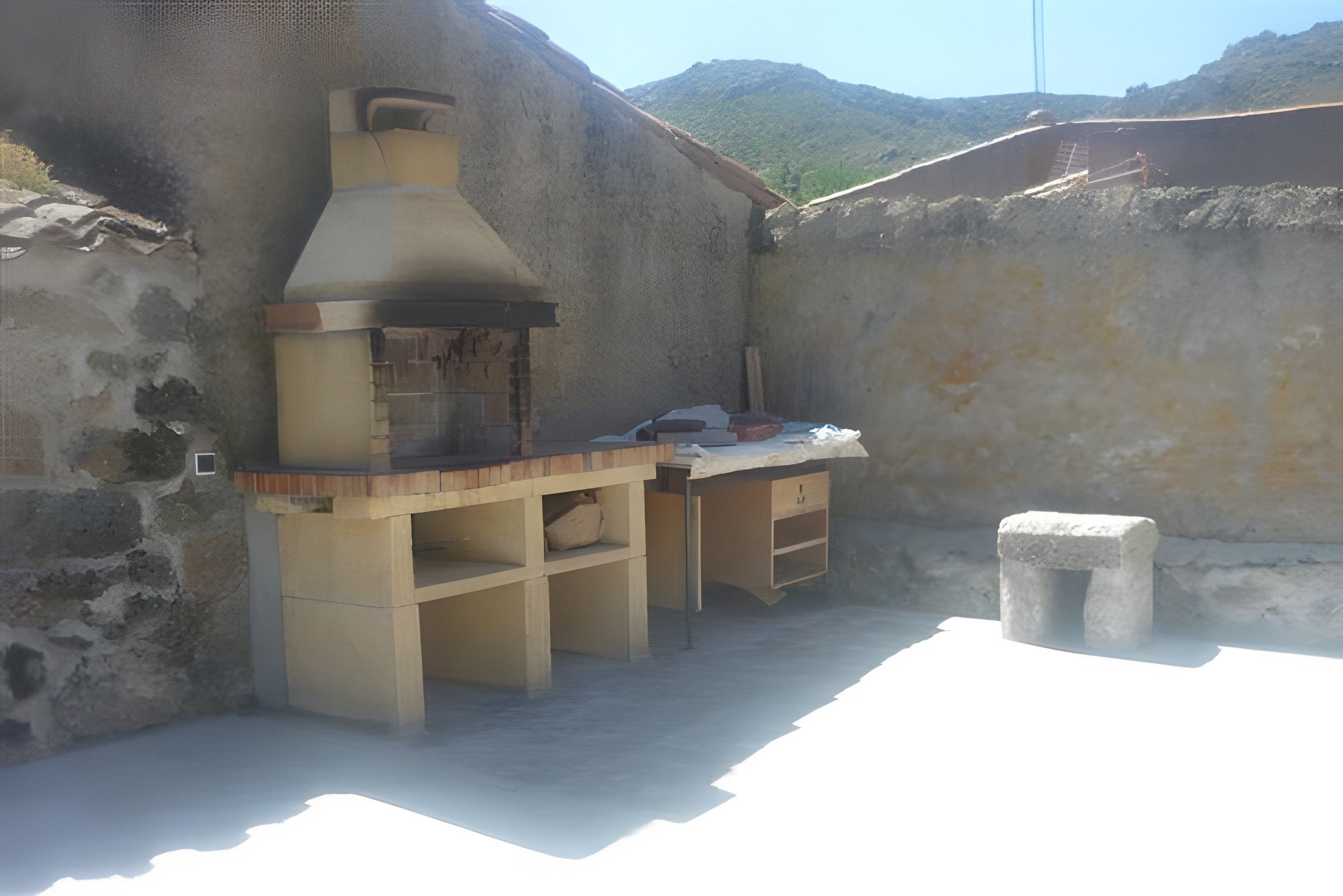 Angolo cucina esterna in pietra con camino, mensole e un piccolo tavolo.