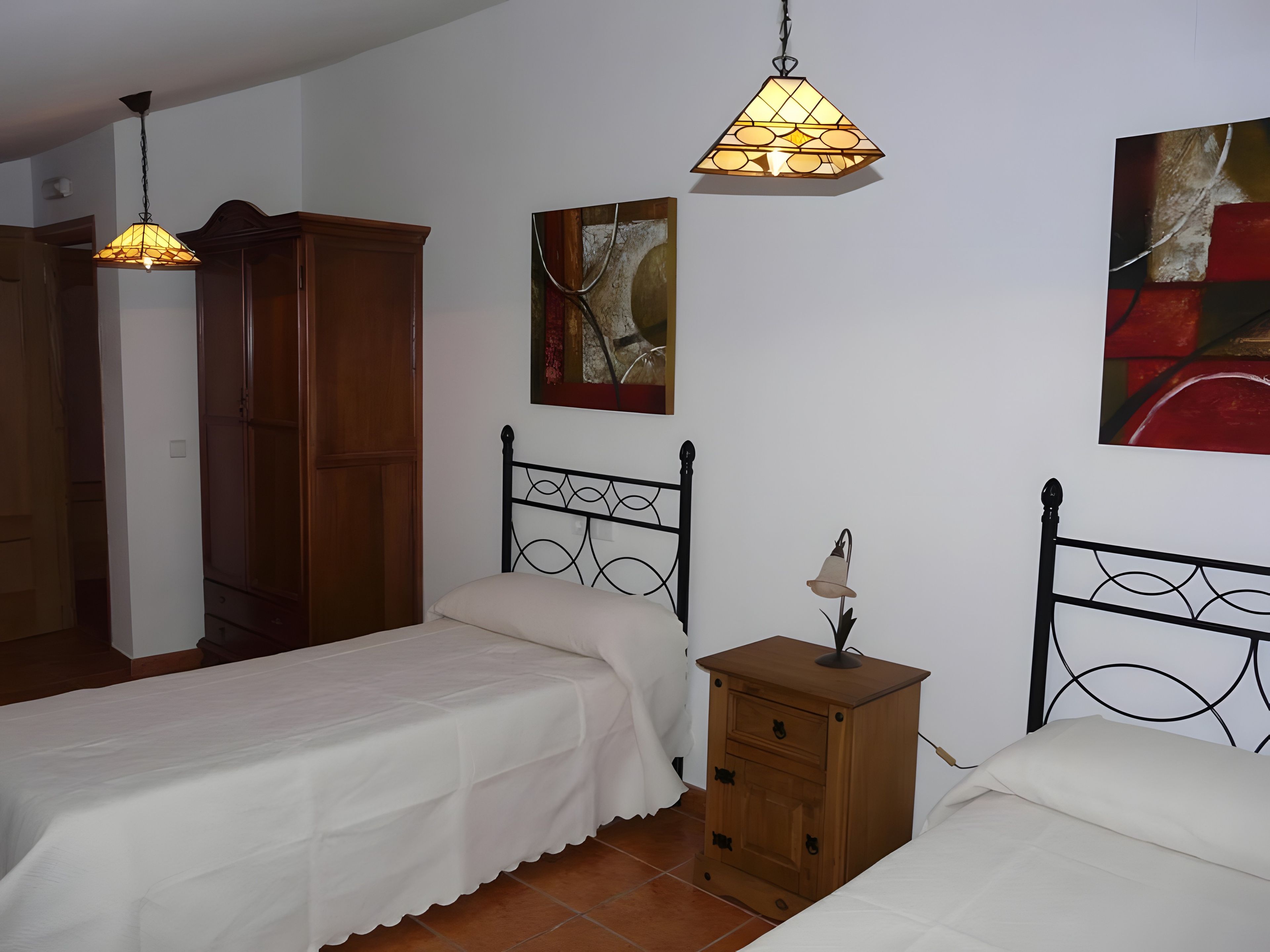 Un dormitorio con dos camas, un armario de madera, una mesita de noche con lámpara y una lámpara colgante.