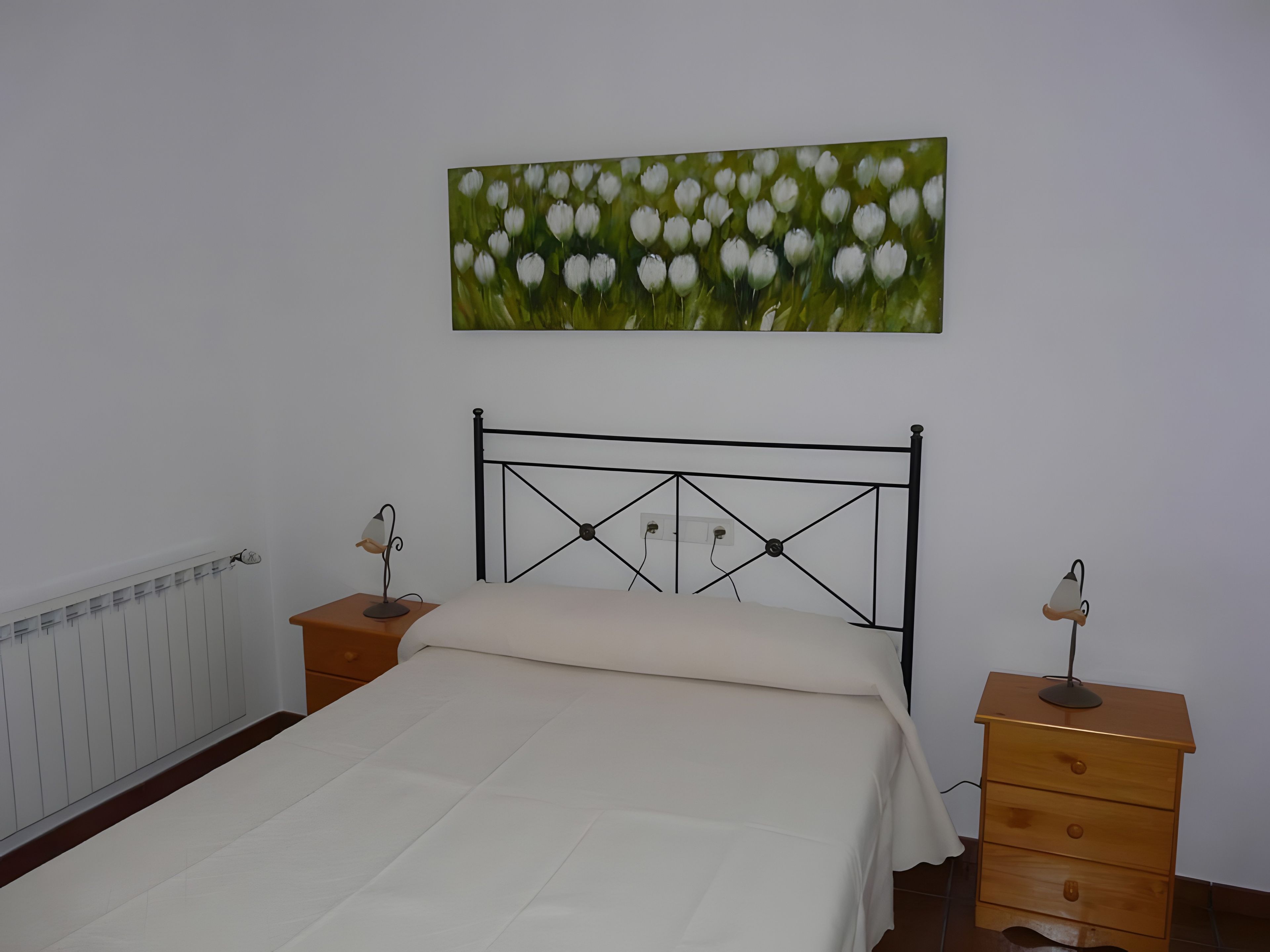 Un dormitorio con cama, dos mesitas de noche, un radiador y un cuadro colgado en la pared.