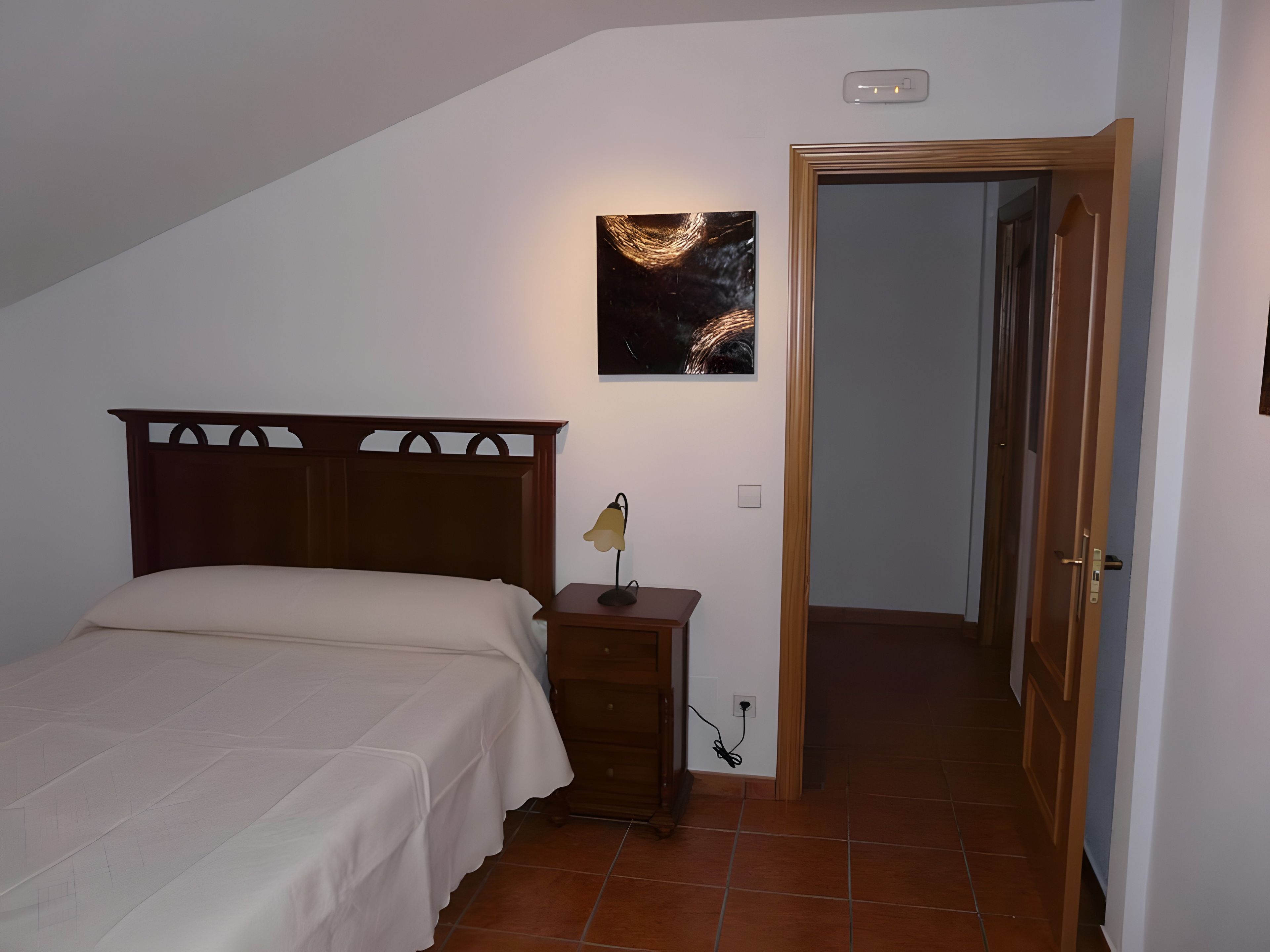 A bedroom with a bed, a nightstand, a wall-mounted lamp, and a door leading to another room.