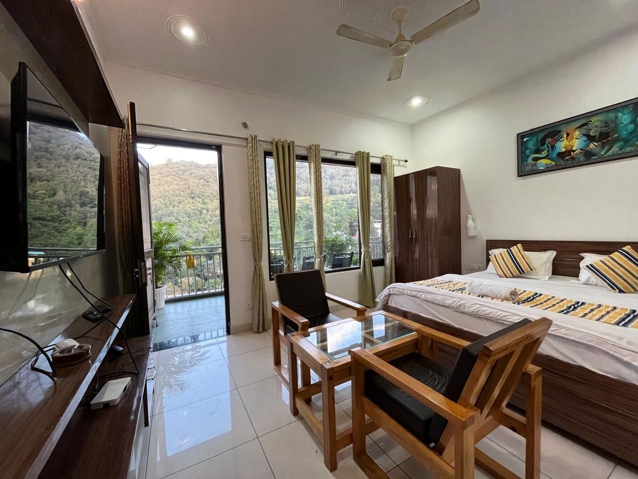 A hotel room with a bed, a TV, a desk with a chair, a balcony with curtains, and a painting on the wall.