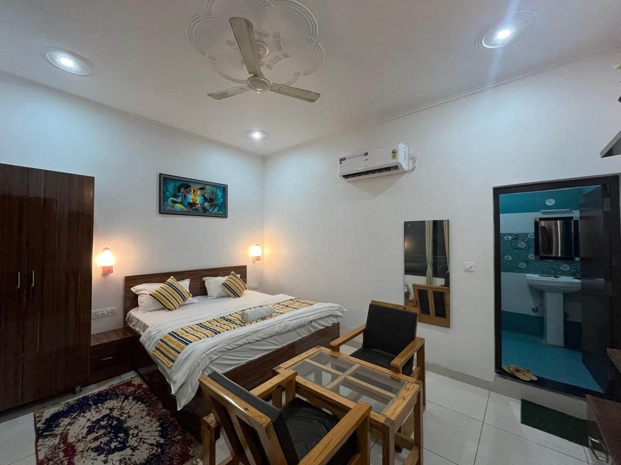 A bedroom with a bed, bedside table, wardrobe, ceiling fan, wall-mounted air conditioner, and a view of the bathroom.